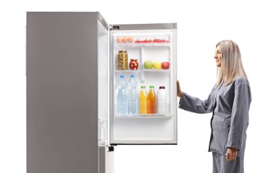 Pijamalı genç bir kadın gecenin bir yarısı beyaz arka planda izole bir buzdolabı açıyor.