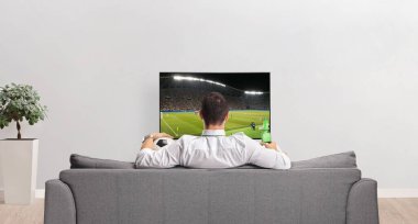 Evde bir şişe bira ve futbol sopasıyla TV ekranının önünde duran bir adamın dikiz görüntüsü.