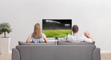 Evde bir kutu patlamış mısırla televizyon karşısında futbol izleyen bir kadın ve bir erkeğin arka plan çekimi.