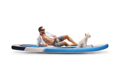 Genç bir adam beyaz arka planda izole edilmiş batılı bir teriyer köpeğiyle SUP tahtasının üzerinde oturuyor.