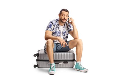 Mutsuz bir adam bavulun üzerinde oturuyor ve beyaz arka planda izole edilmiş bir akıllı telefon kullanıyor.