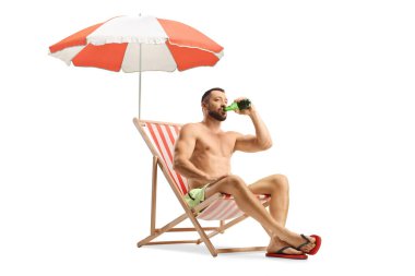 Mayolu genç adam beyaz arka planda izole edilmiş şemsiyenin altında plaj sandalyesinde oturmuş bira içiyor.