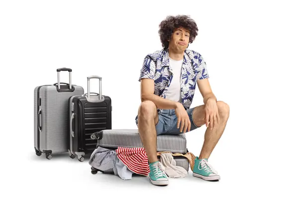 stock image Tired young man sitting on a suitcase full of clothes isolated on white background