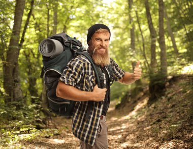 Sırt çantalı bir yürüyüşçü ormanda gülümsüyor ve baş parmağını kaldırıyor.