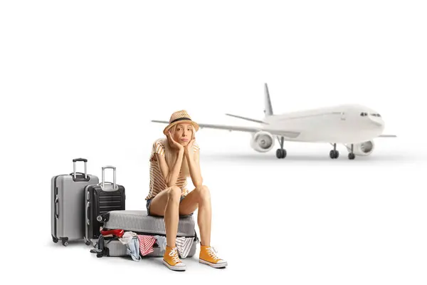 stock image Confused female toursit sitting on a suitcase in front of an airplane isolated on white background