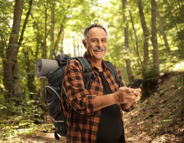 Sırt çantasıyla ormanda akıllı telefon tutan ve gülümseyen olgun bir yürüyüşçü.
