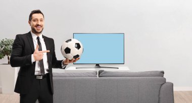 Evde elinde futbol topu tutan, kanepe ve televizyonu işaret eden profesyonel bir adam.