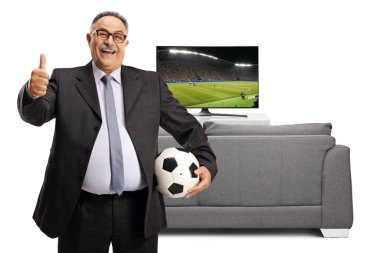 Bir futbol topu tutan heyecanlı olgun bir adam ve beyaz arka planda izole edilmiş bir kanepe ve televizyon ekranının önünde işaret eden bir başparmak.