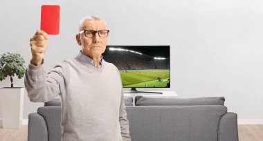 Ciddi yaşlı bir adam TV ekranının önünde kırmızı kart gösteriyor. Oturma odasında futbol maçı var.