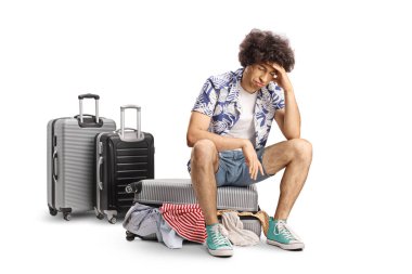 Aşırı toplanmış bavulun üzerinde oturan üzgün genç adam beyaz arka planda izole edilmiş.