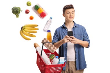 Akıllı telefonlu genç bir adam elinde alışveriş sepetiyle sepetin içine düşen ürünleri beyaz arka planda izole bir şekilde tutuyor.