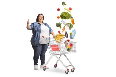 Elinde alışveriş arabası olan şişman bir kadın baş parmak işareti yapıyor ve beyaz arka planda yiyecek alışverişi yapıyor.