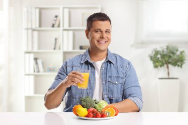 Elinde bir bardak portakal suyu ve bir tabak meyve ve sebzeyle evdeki masada oturan genç adam.