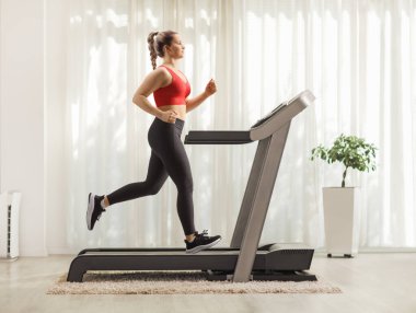 Evdeki koşu bandında koşan güçlü genç bir kadının tam boy profil fotoğrafı.