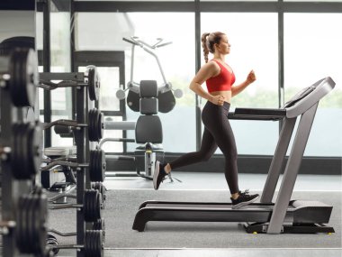 Spor salonundaki koşu bandında koşan güçlü genç bir kadının tam boy profil fotoğrafı.