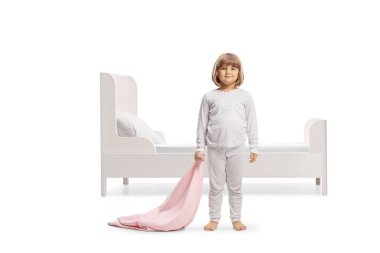 Beyaz arka planda izole edilmiş bir yatağın önünde pembe bir battaniye tutan pijamalı bir çocuğun tam boy portresi.