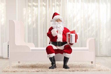 Noel Baba bir odada bir yatakta oturuyor ve elinde bir hediye tutuyor.