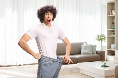 Şok olmuş zayıf bir adam kot pantolon deniyor ve evde gülümsüyor.