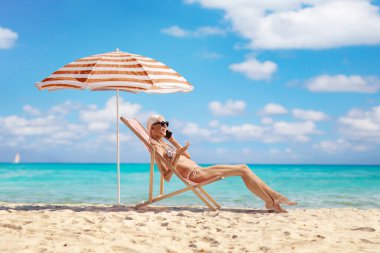 Plaj sandalyesinde oturan ve deniz kenarında cep telefonu kullanan bikinili mutlu bir kadın.