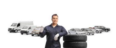 Üniformalı bir otomobil tamircisi beyaz arka planda izole edilmiş bir otoparkta lastik yığınına yaslanıyor.