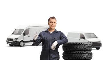 Auto mechanic worker with vans leaning on a pile of tires and showing a thumb up gesture isolated on white background clipart