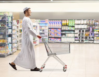 Full length profile shot of a young muslim man walking and pushing a shopping cart at a supermarket clipart