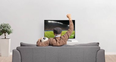 Kanepede oturan yaşlı bir adamın elinde futbol topu ve kanepede televizyon karşısında tezahürat yapması. 