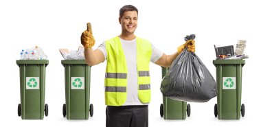 Siyah plastik bir torba tutan atık toplayıcı ve beyaz arkaplanda izole edilmiş geri dönüşüm konteynırlarının önünde baş parmaklarını gösteriyor