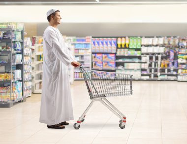 Geleneksel Müslüman cüppeli bir adamın süpermarkette bir alışveriş arabasını iterken tam boy profil fotoğrafı.