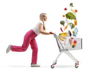 Beyaz arka planda izole edilmiş alışveriş arabasında alışveriş yapan genç bir kadının tam boy profil çekimi.