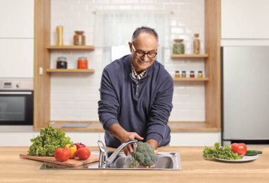 Evdeki mutfak lavabosunda sebze yıkayan bir adam.