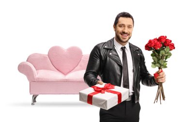 Deri ceketli bir adam bir hediye kutusu ve beyaz arka planda izole edilmiş pembe kadife bir kanepenin önünde bir demet kırmızı gül tutuyor.