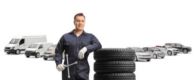 Üniformalı bir otomobil tamircisi beyaz arka planda izole edilmiş bir otoparkta lastik yığınına yaslanıyor.