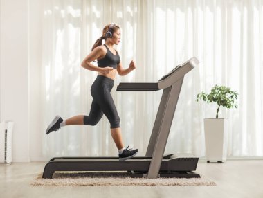 Evdeki koşu bandında kulaklık takan Asyalı genç bir kadının tam boy profil fotoğrafı.