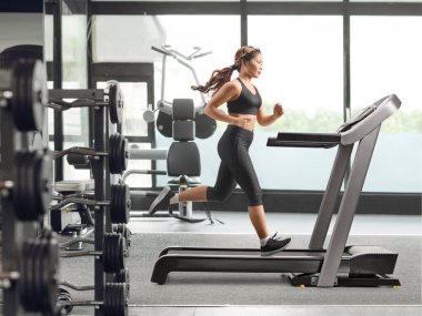 Spor salonunda koşu bandında koşan Asyalı bir kadının tam boy profil fotoğrafı.
