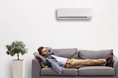 Beyaz arka planda izole edilmiş bir havalandırma ünitesinin altındaki kanepede dinlenen adam.