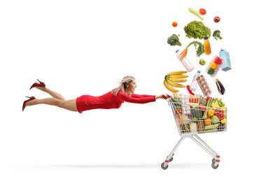 Kırmızı elbiseli çekici bir kadın beyaz arka planda izole edilmiş alışveriş arabasıyla uçuyor ve elinde alışveriş arabası tutuyor.