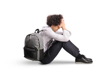Sırt çantalı üzgün bir erkek öğrenci beyaz arka planda izole bir şekilde yerde oturuyor.