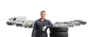 Otomotiv işçisi üniformalı bir araba parkında duruyor ve beyaz arka planda izole edilmiş bir lastik yığınına yaslanıyor.