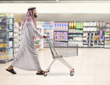 Suudi Arabistan 'lı bir adamın süpermarkette yürürken ve alışveriş arabasını iterken tam boy profil fotoğrafı.