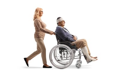 Beyaz arka planda kafası, kolu ve bacağı yaralı bir adamı tekerlekli sandalyeye iten kadın. 