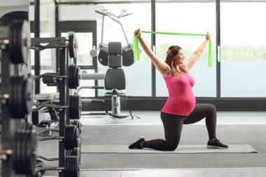 Hamile bir kadın spor salonunda diz çöküp bantla egzersiz yapıyor.