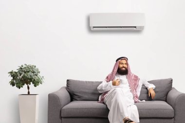 Saudi arab man sitting on a sofa with a cup of tea under an air conditioner clipart