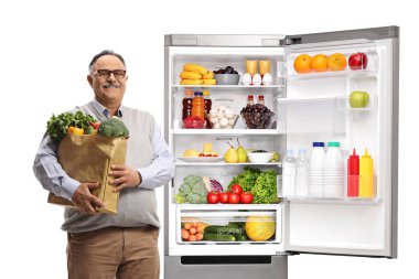 Bakkal çantalı olgun bir adam beyaz arka planda izole edilmiş açık bir buzdolabının yanında duruyor.