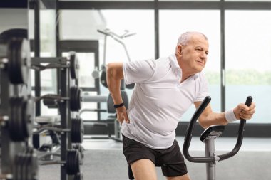 Spor salonunda bisiklet süren, sırt çantalı, olgun bir adam.