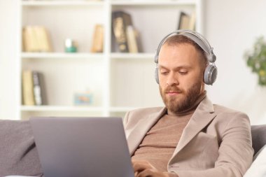 Kulaklıklı bir adam evde dizüstü bilgisayar kullanıyor.