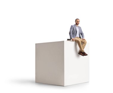 Beyaz arka planda beyaz bir küpün üzerinde oturan kitaplı adam.