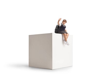 Üniformalı kız öğrenci beyaz bir kübün üzerinde oturuyor ve beyaz arka planda izole bir şekilde el sallıyor.