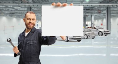 Araba garajında İngiliz anahtarı ve küçük boş bir kart tutan üniformalı bir tamirci.