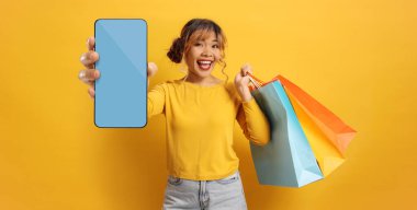 Alışveriş torbaları taşıyan mutlu Asyalı kadın ve sarı arka planda akıllı telefon.